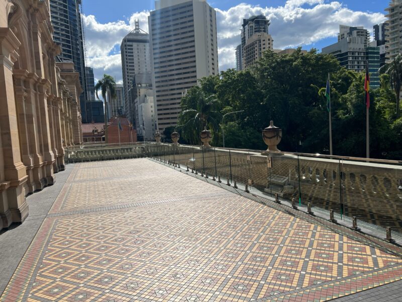 Parliament House balcony with heritage encaustic tiles and new frameless glass balustrade, Brisbane CBD towers behind.