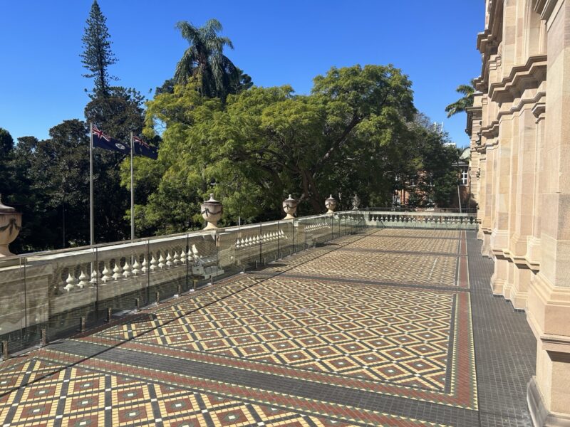 Open balcony terrace with tiled floor, glass balustrade and the Parliament gardens and flags beyond.