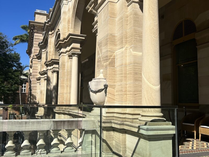 Glass balustrade returning around a sandstone pier and urn at the balcony corner.