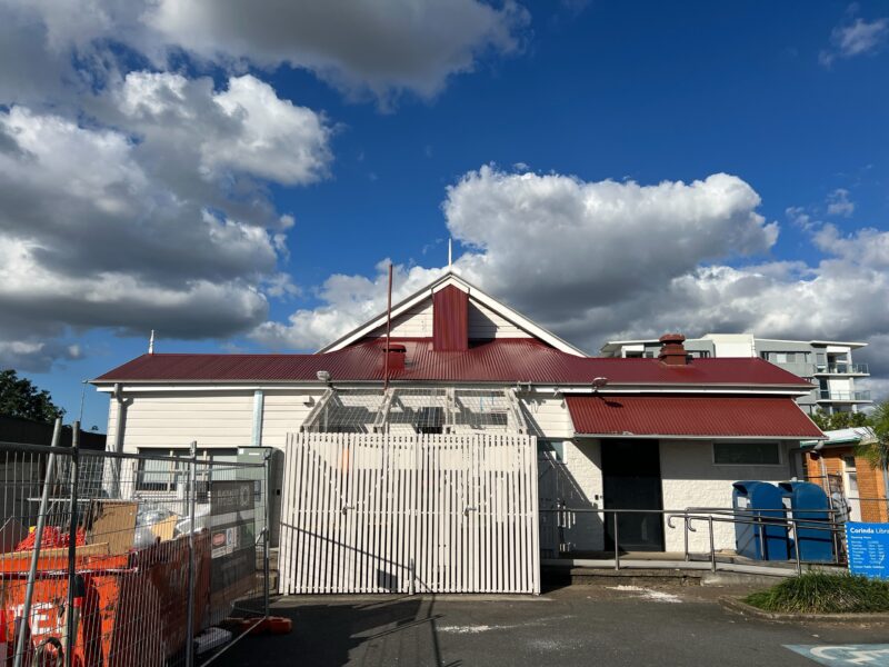 Rear of the library with red roof under a cloudy blue sky, new screened plant enclosure and site fencing.