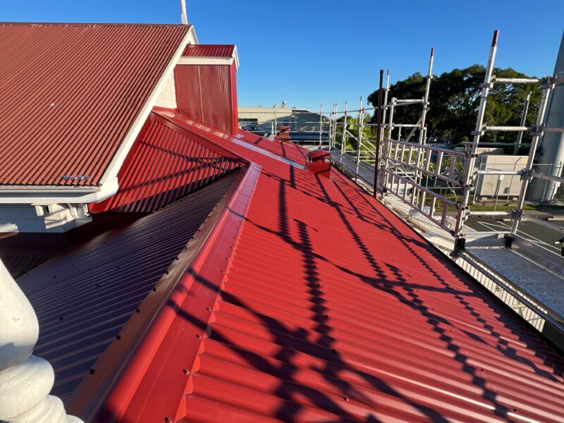 Close view of red roof sheeting and ridge capping with scaffold shadows.
