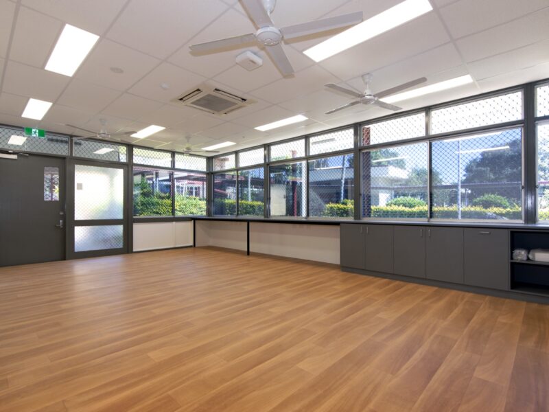 Large open-plan staff room with timber-look vinyl floor, full-width windows, ceiling fans and grey joinery along the window line.