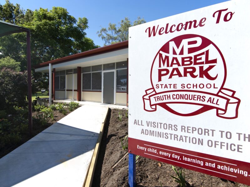 Mabel Park State School 'Welcome To' sign in the foreground with the refurbished administration building, new concrete path and garden beds under blue sky.