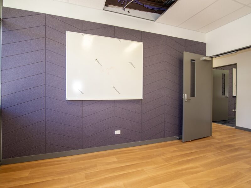 Meeting room with purple herringbone acoustic wall, whiteboard, grey door and timber-look floor.