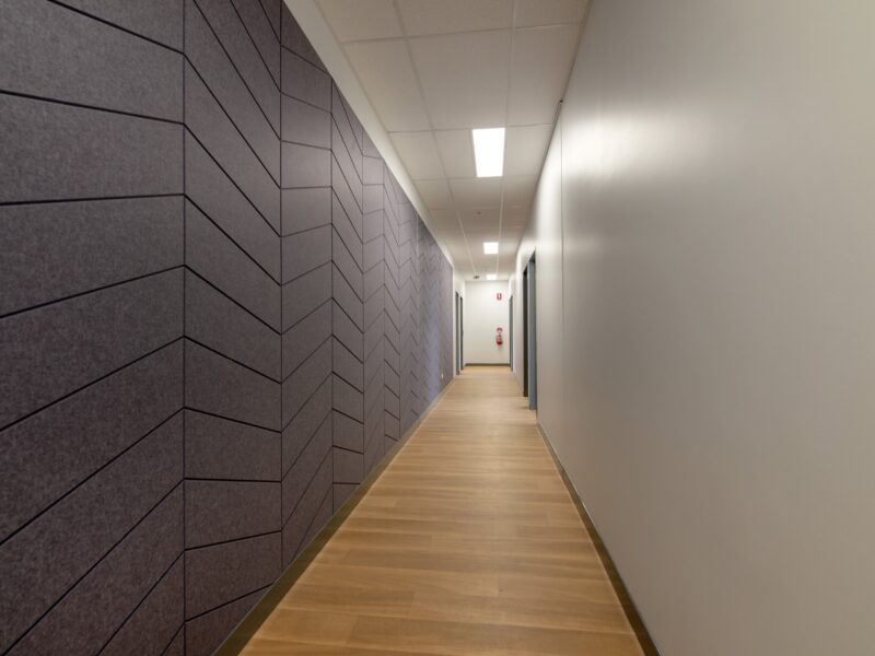 Long corridor with herringbone-patterned purple acoustic wall panels on one side, timber-look floor and linear ceiling lights.