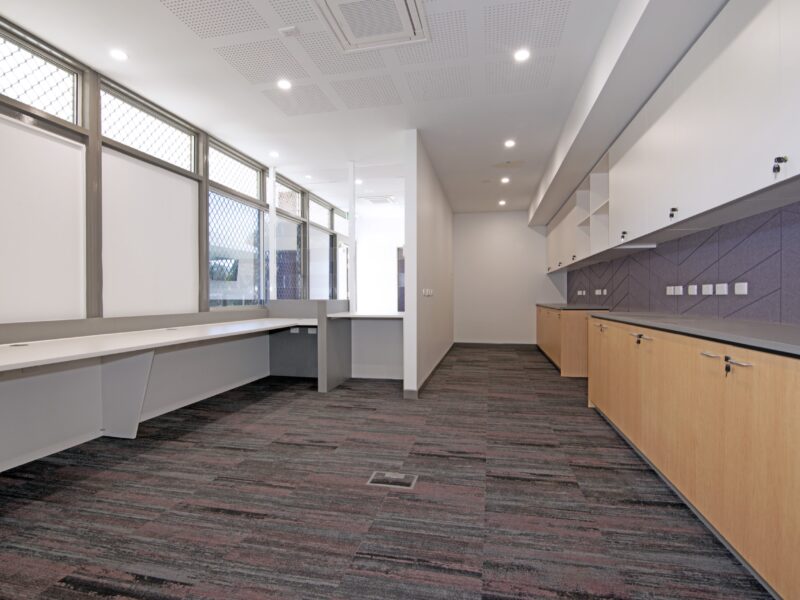 Administration workroom with carpet tiles, perimeter desk under windows and blonde-timber base cabinets with overhead white cupboards.