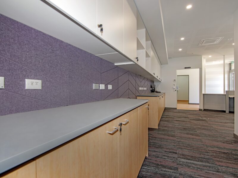 Close angle along grey benchtop and timber cabinetry against a purple acoustic-panel splashback with power outlets.