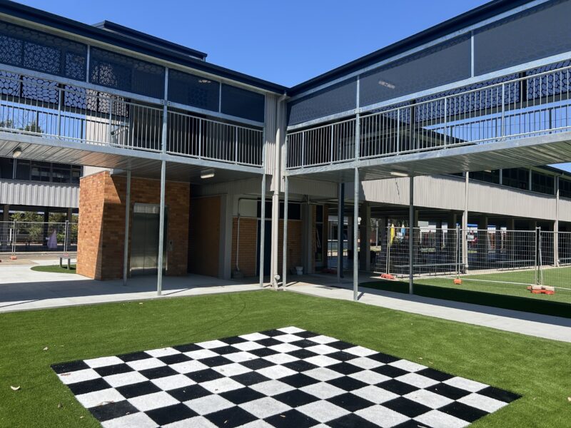 School courtyard with new two-storey walkways, perforated screens, lift and chequerboard artificial turf.