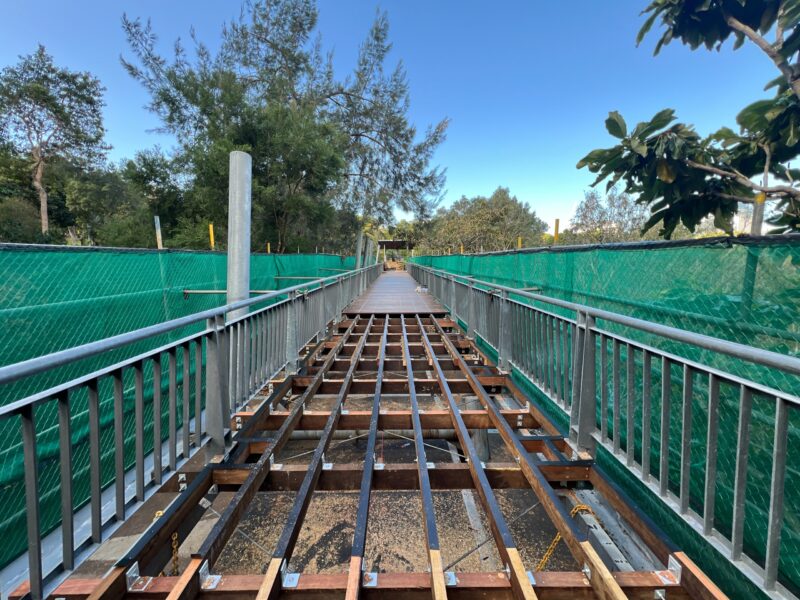 Deck boards removed exposing new timber joists and bearers along the footbridge, with green shade-cloth on the balustrades.