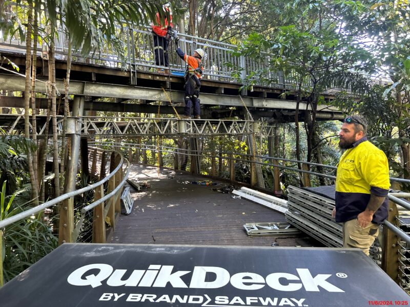 Crew working at height on the elevated footbridge above QuikDeck suspended scaffold panels, with a site worker in hi-vis in the foreground.