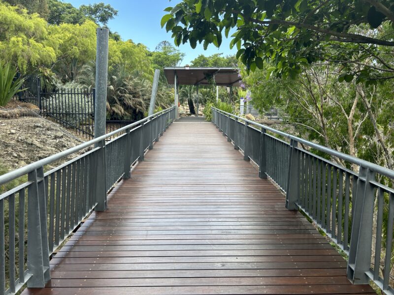 Footbridge with new decking leading to a shade-roofed lookout, framed by trees and blue sky.