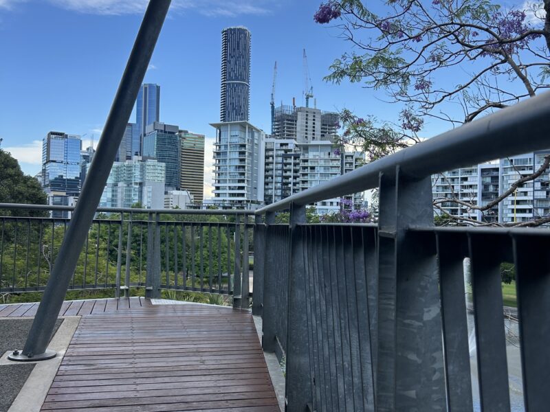 New decking at a bridge corner with the Brisbane CBD skyline and jacaranda blooms beyond.