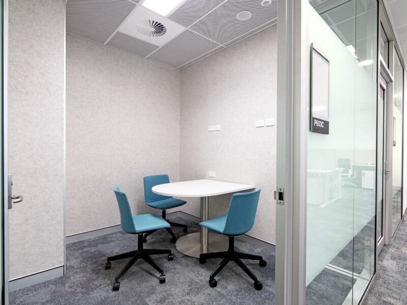 Small meeting room with round table and teal chairs behind glass partition.