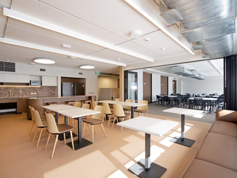 Sunlit staff breakout area with white tables, timber chairs, leather banquette and exposed spiral ductwork opening onto a learning space.