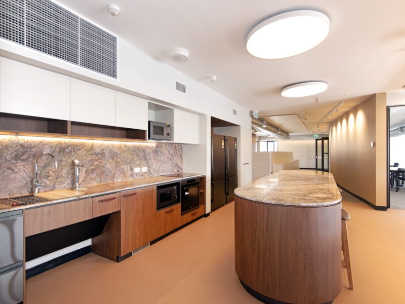 Fitted kitchenette with stone splashback, timber joinery and curved stone-topped island bench.