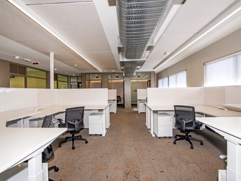 Open-plan workstation area with white sit-stand desks, black task chairs and exposed ductwork overhead.