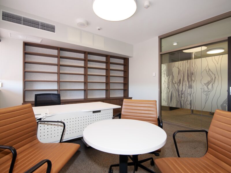 Academic office with tan leather meeting chairs, round table, desk and full-height timber shelving.