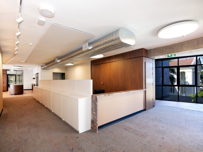Reception area with white counter, timber feature wall, track lighting and exposed duct leading to a glazed exit.