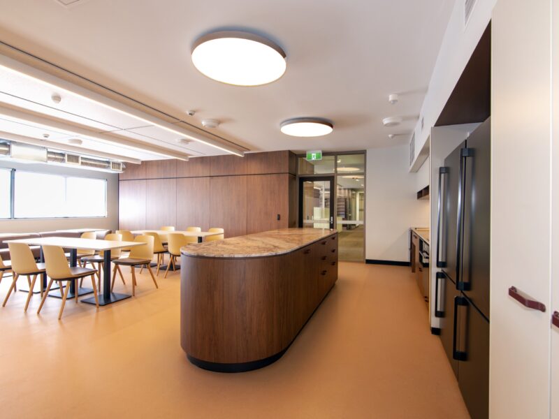 Curved timber island bench with stone top in the breakout kitchen, dining tables beyond.