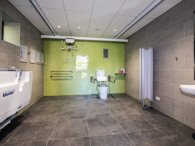 Changing Places accessible bathroom with green tiled wall, ceiling hoist, change table and grey floor tiles.