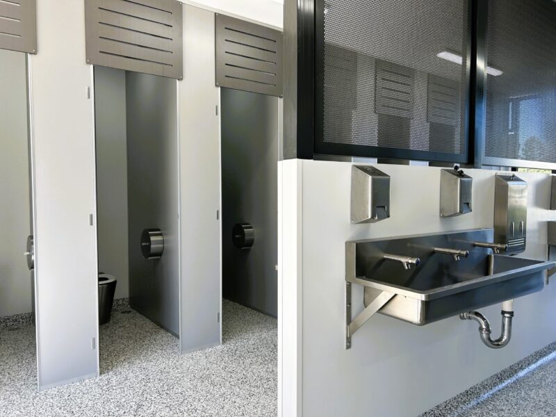 Full-height toilet partitions beside a stainless steel trough basin with soap and towel dispensers.